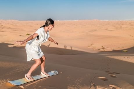 A woman in a white dress sandboards down a vast, sunny desert dune under a clear blue sky, conveying a sense of adventure and freedom.