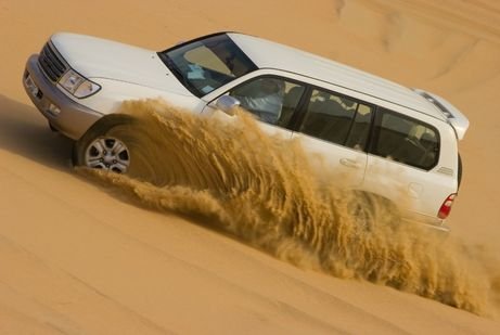 A white SUV in UAE drives through the desert, kicking up a dramatic spray of sand. The vehicle tilts sideways on a steep dune, suggesting adventure.