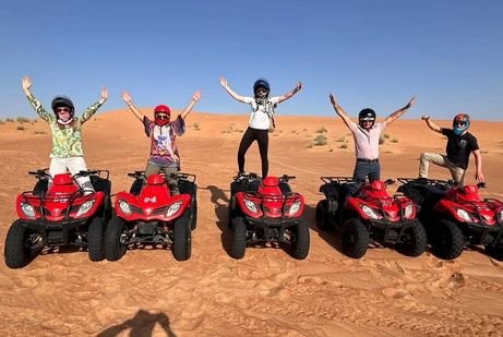 Five people on red ATVs raise their arms excitedly in a UAE desert landscape, under a clear blue sky, conveying a sense of adventure and joy.