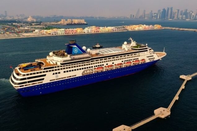 An Arabian cruise ship with a blue hull sails in a sunlit harbor, with a Dubai City skyline and colorful waterfront buildings in the background.