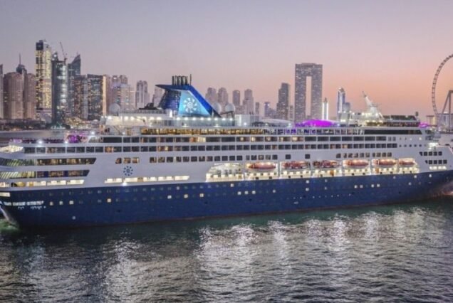 Arabian Cruise ship docked in a Dubai vibrant harbor, with a city's illuminated skyline at dusk. The scene is calm, reflecting lights on the water.