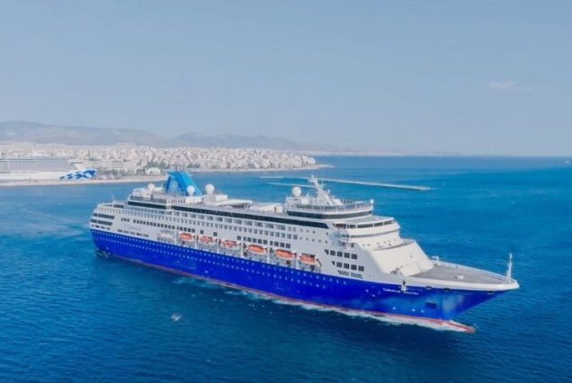 A large blue and white Arabian cruise ship sails through calm, clear blue waters near a distant coastal city. The sky is clear, conveying a bright and peaceful atmosphere.