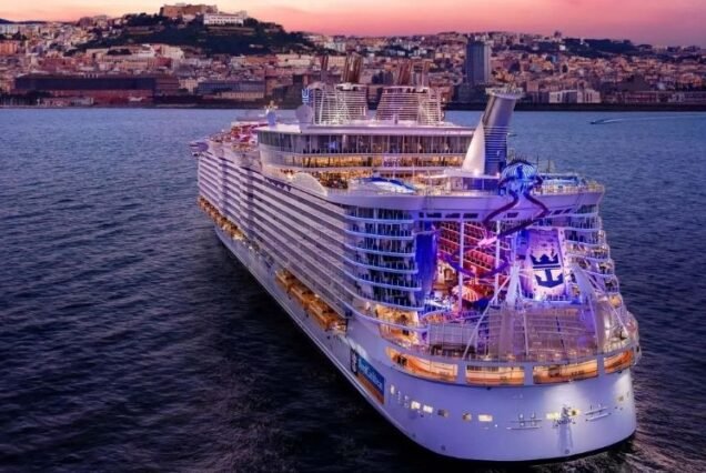 A large, Royal Caribbean Cruises well-lit cruise ship sails at sunset with a scenic cityscape on the horizon. The colorful lights create a festive ambiance on the water.