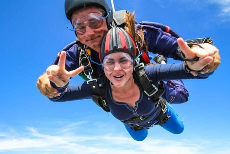 Skydiving in tandem against a bright blue sky in Dubai. Both are smiling, making peace signs, and wearing safety gear.