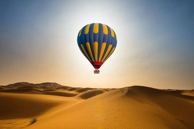 A vibrant blue and yellow hot air balloon soars over vast, sunlit desert dunes under a clear sky, evoking a sense of adventure and tranquility.