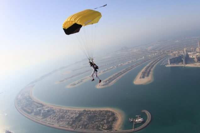 Skydiver with a yellow parachute descends over Dubai's Palm Jumeirah, a man-made island chain, with the cityscape and ocean in the background.