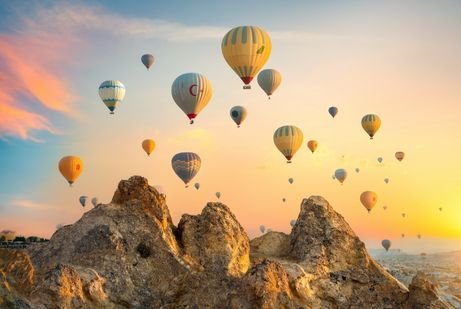 A serene scene of numerous hot air balloons floating over rocky terrain at sunrise in Dubai. The sky is filled with warm orange and pink hues, creating a peaceful atmosphere