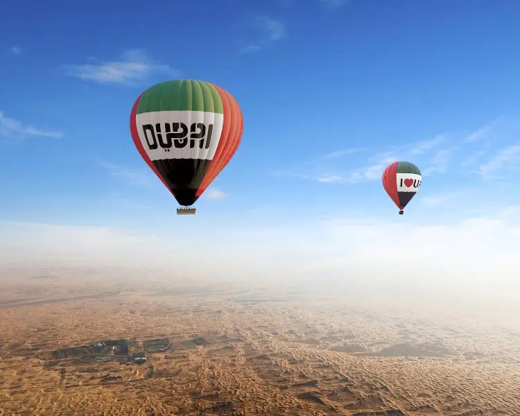 Colorful hot air balloon flying over Dubai desert at sunrise, with golden sand dunes and the city skyline in the distance.